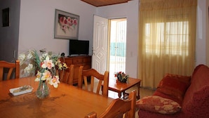 Un comedor con una mesa de madera, sillas, un florero, un televisor sobre un mueble bajo y una puerta que da al exterior.