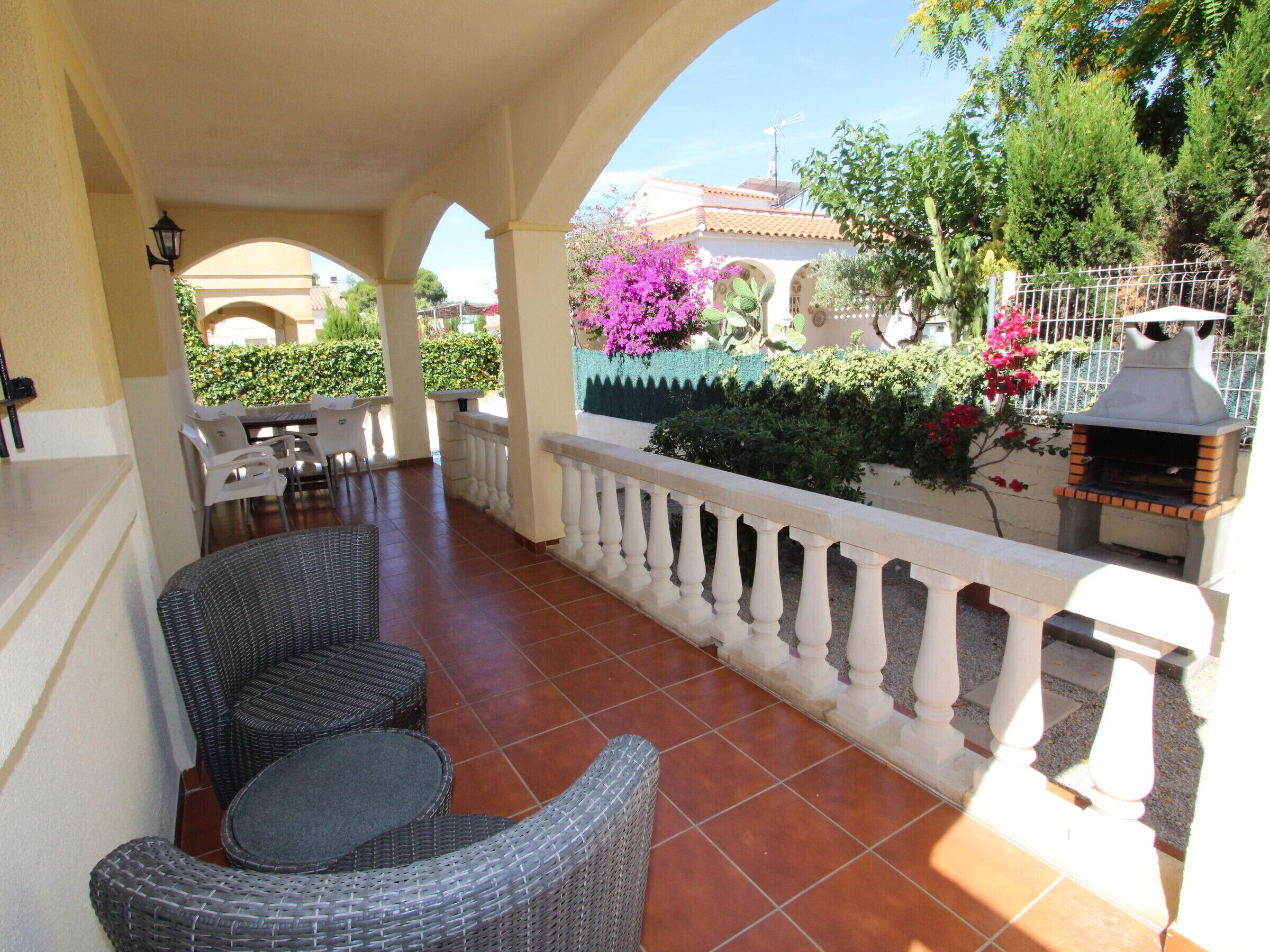 A covered patio area with wicker furniture, a barbecue grill, and a view of a building with a tiled roof.