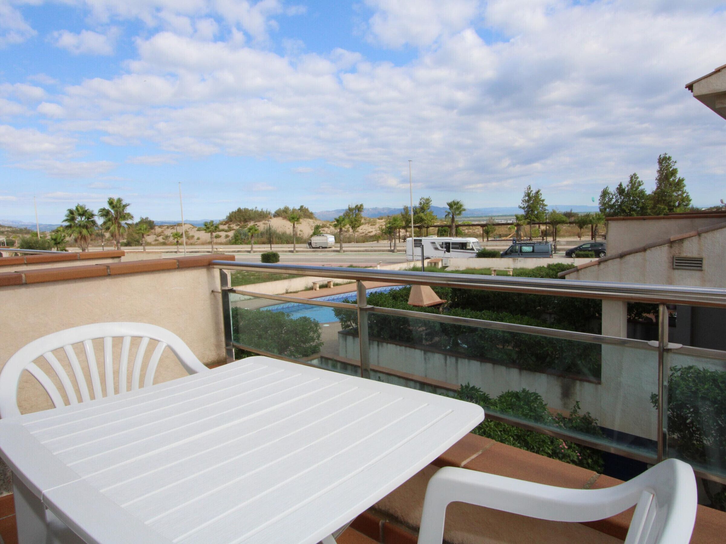 A balcony with a table and chairs, overlooking a parking area with vehicles and a pool.