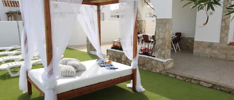 A four-poster bed with white linens and a canopy, set on artificial grass, with a stone wall and outdoor furniture in the background.