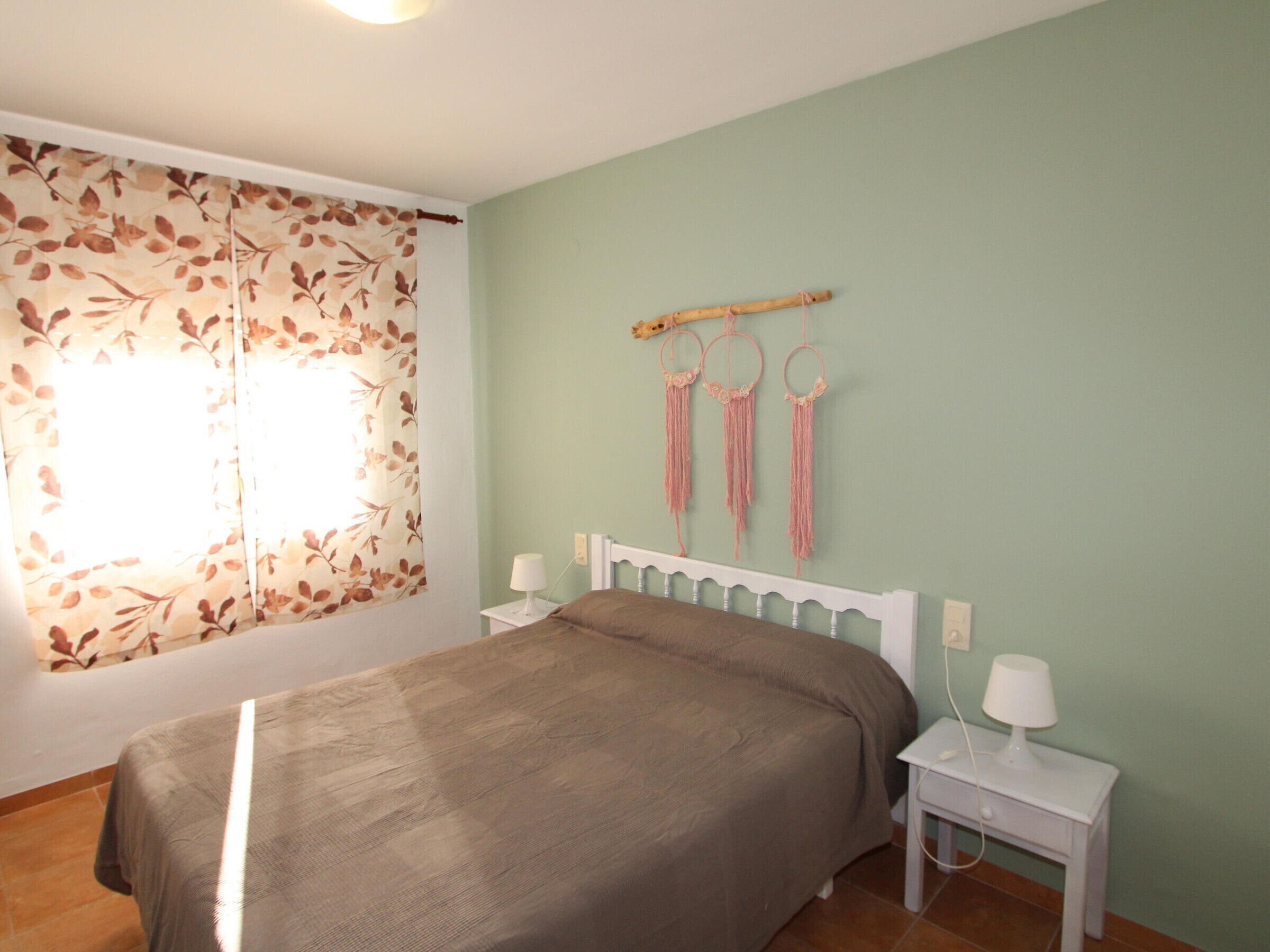 Ein Schlafzimmer mit Bett, einem Fenster mit floralem Vorhang, einem Nachttisch mit Lampe und einem an der Wand hängenden Traumfänger.