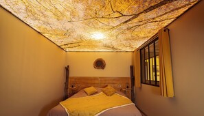 A room with a bed, a wooden headboard, a wall-mounted circular object, and a window with curtains. - Gite de Rochebrune, Évasion et Déconnexion au Cœur des Monts du Cantal (Neuvéglise-sur-Truyère)