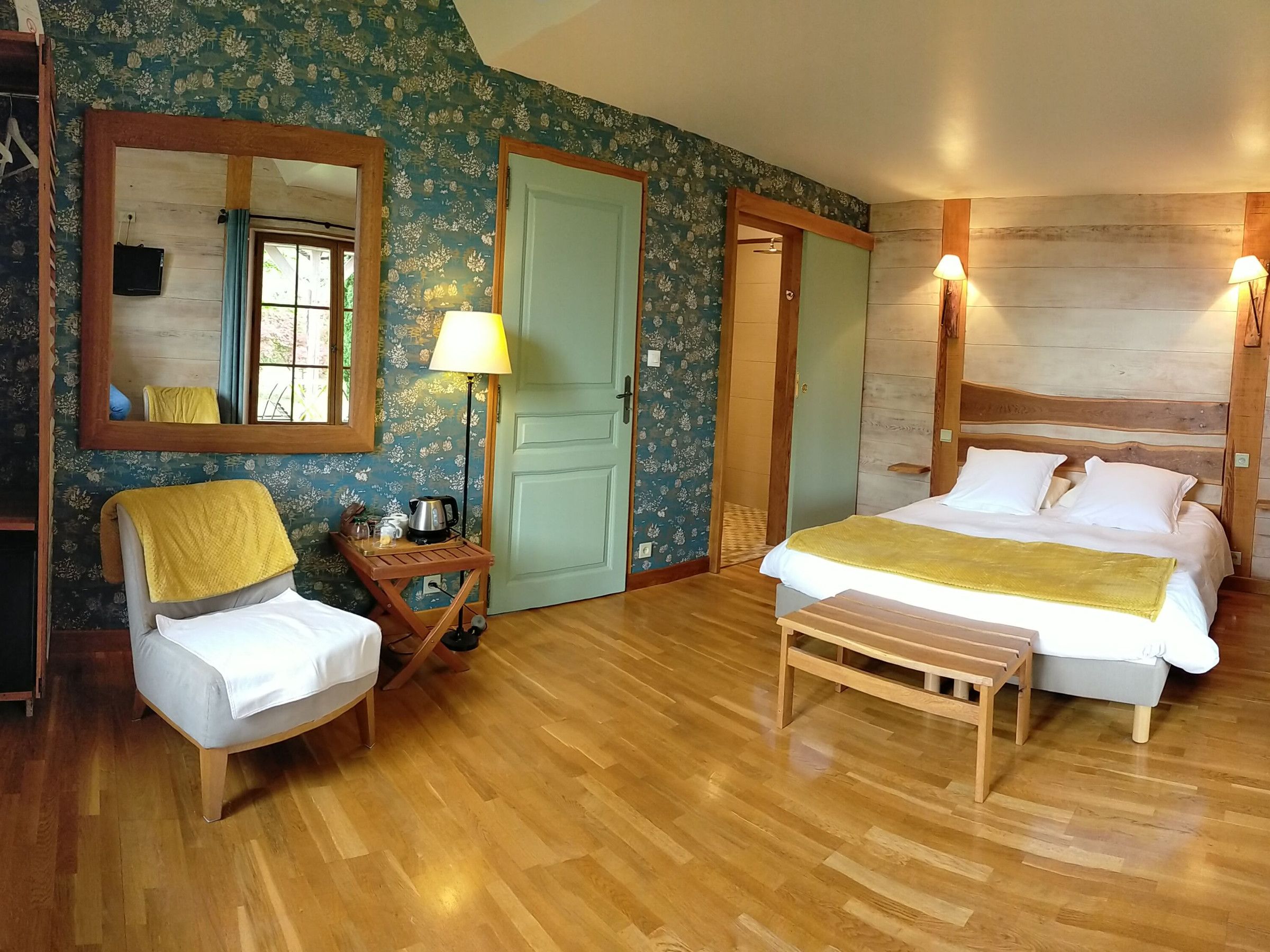 Un dormitorio con cama de madera, una mesita de noche con lámpara, una silla con toalla, un espejo y una puerta.