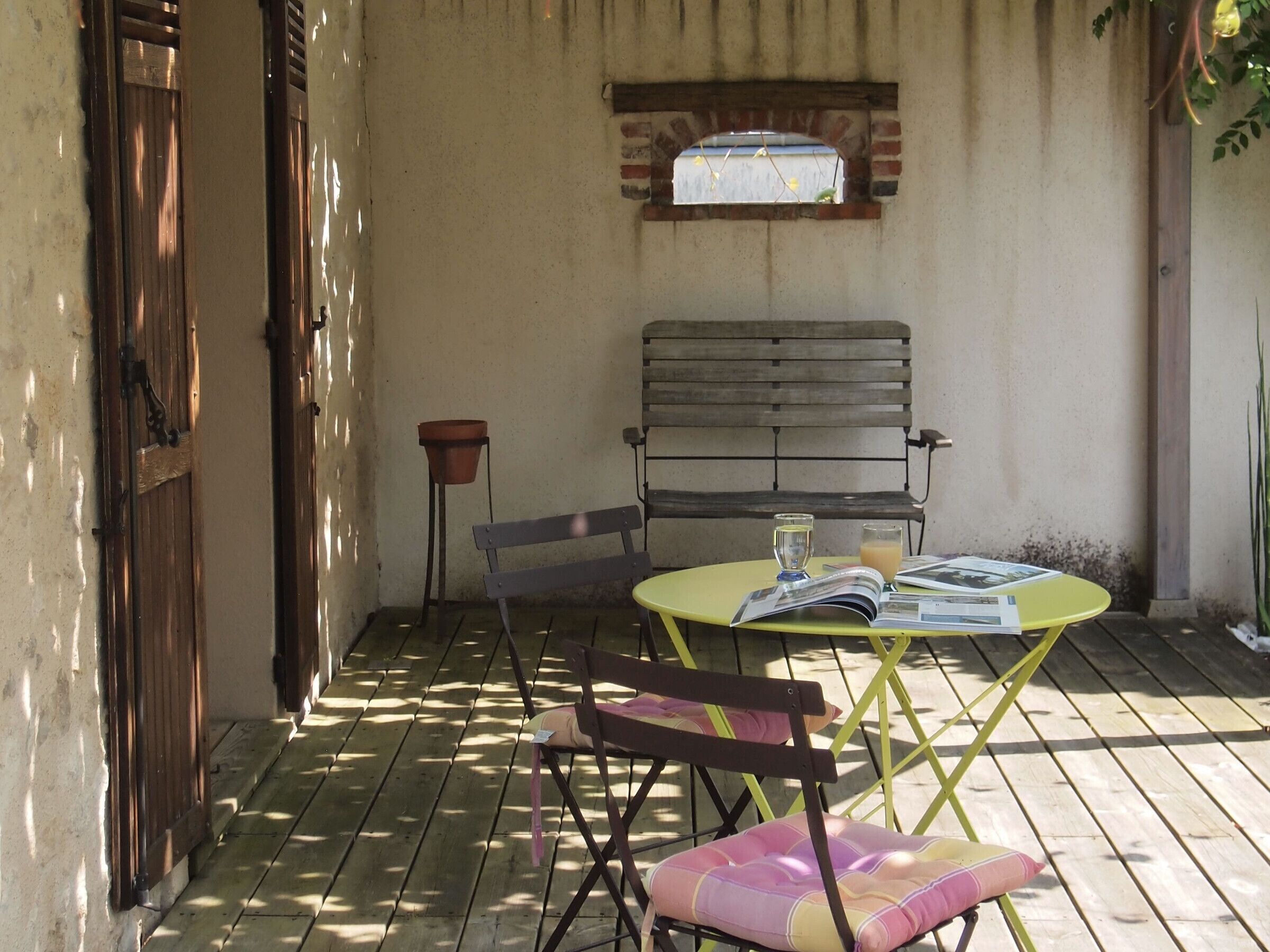 An outdoor patio with a wooden floor, a round yellow table, two chairs, a bench, and a small window with a view.