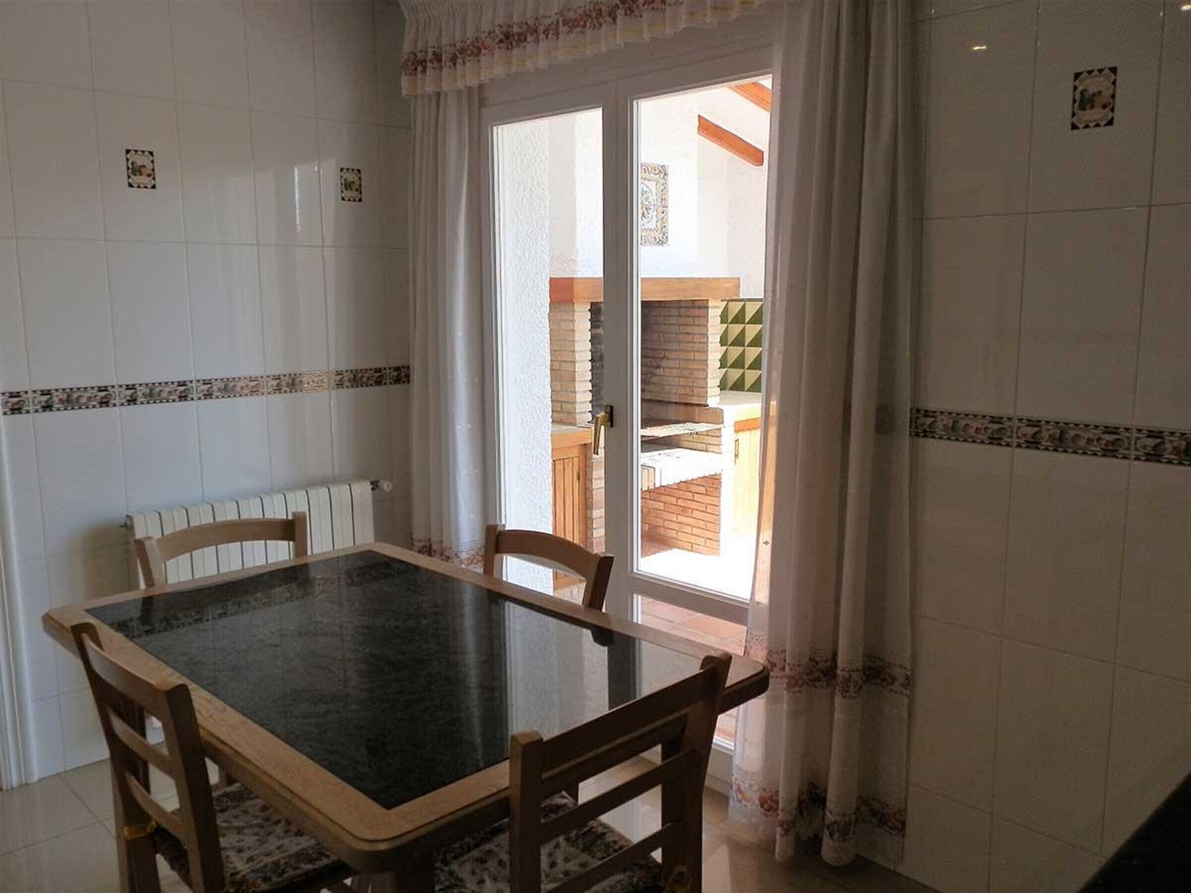 Een eethoek met een tafel en stoelen, een raam met gordijnen en uitzicht op een balkon met een houten hekwerk.