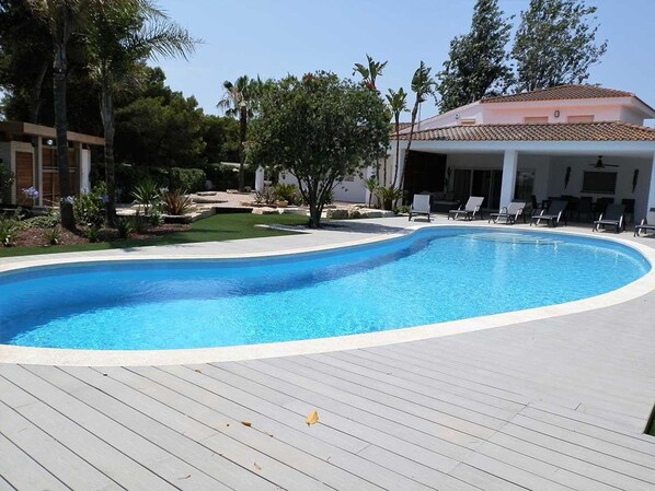 A swimming pool with a wooden deck, surrounded by palm trees and a building with a tiled roof. - Cozy Beachside House: Private Pool, Free WiFi, Pet-Friendly, Near Dog Beach (Deltebre)