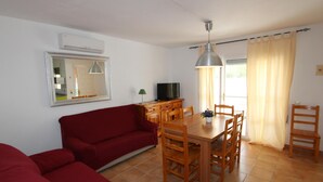 A compact living space with a red sofa, wooden dining table, and chairs. A television is on a wooden cabinet, and a mirror hangs on the wall. - Cozy Beachside Home: Private Pool, Pet-Friendly, WiFi, A/C Option, Near Dog Beach (Deltebre)