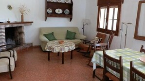 Un salon chaleureux avec une cheminée en briques, un canapé, une table à manger avec des chaises et une bibliothèque ornée d’assiettes décoratives.