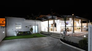 Ein modernes Haus mit überdachter Terrasse, einem kleinen Rasen und einer Sitzecke mit Kissen.