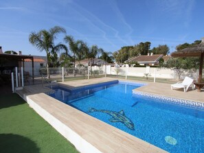 A swimming pool with a dolphin mosaic, surrounded by a fence, with a grassy area and houses in the background. - Family Haven Near the Sea: Private Pool, Pet-Friendly, Wi-Fi, Optional A/C (Deltebre)