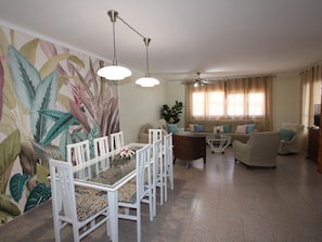 Un coin repas avec une table en verre et des chaises blanches, un salon avec un canapé et des fauteuils, et un ventilateur de plafond.