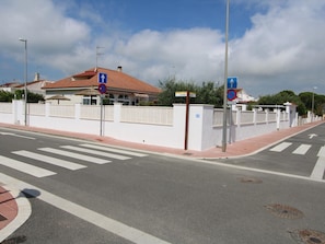 A street intersection with a pedestrian crossing, a white wall with a fence, and a building with a red roof.