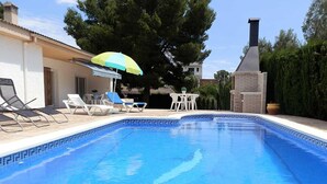 Una piscina con sillas de descanso y una mesa, rodeada de follaje y un edificio con chimenea.