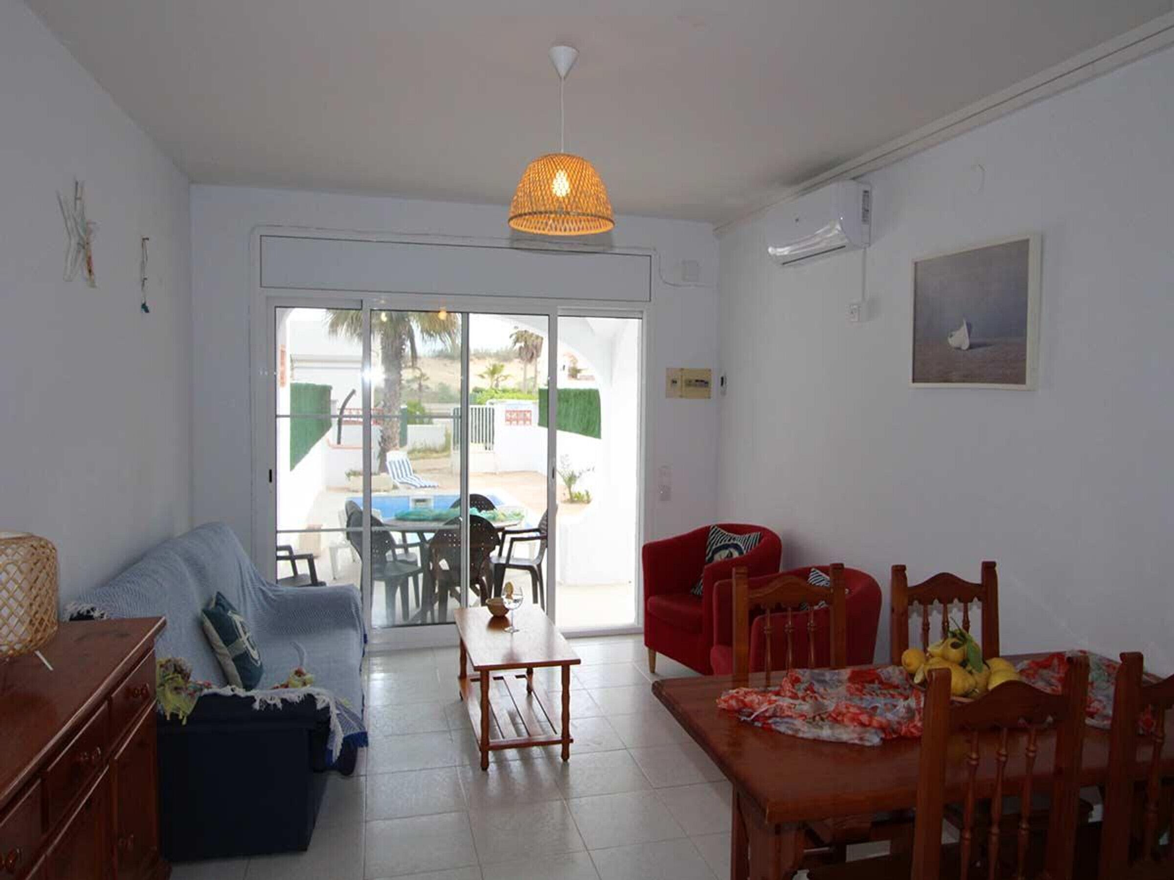 Un salon comprenant un canapé, une table à manger avec des chaises, un luminaire suspendu et une porte-fenêtre coulissante donnant sur un espace extérieur avec une piscine et des chaises.