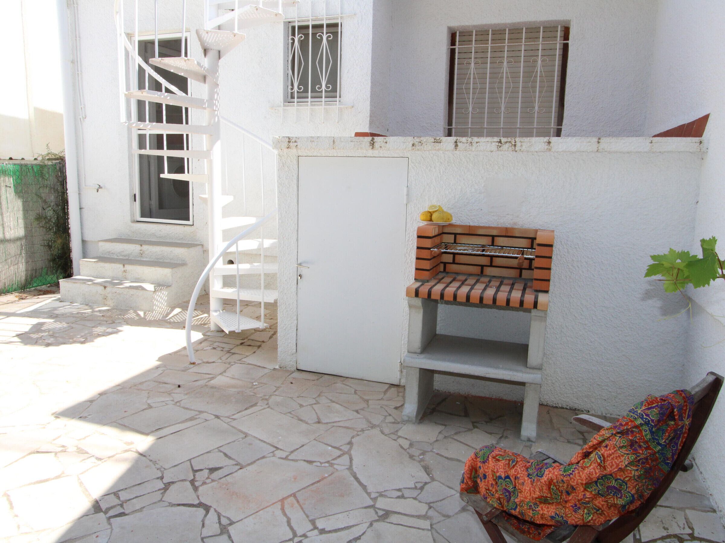 A patio area with a white spiral staircase, a brick barbecue grill, a bench, and a chair with a colorful cover.