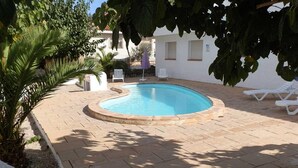 Una piscina con borde curvo, rodeada por un patio empedrado. Hay sillones de descanso y una mesita cerca de la piscina.