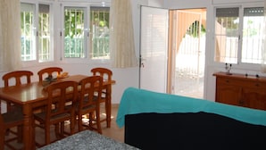 Een kamer met een eettafel en stoelen, een bank, een raam met gordijnen, en een deur die naar buiten leidt.