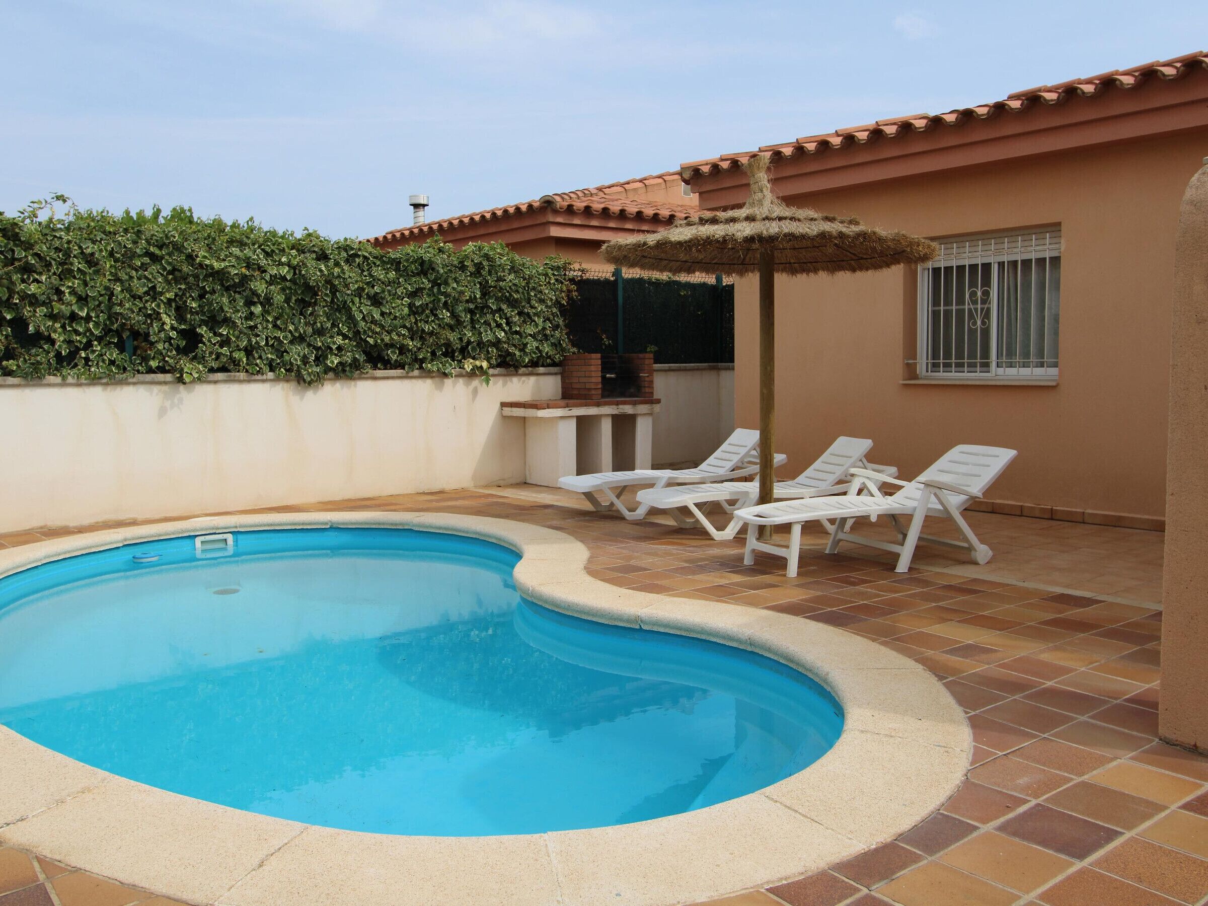 Une piscine bordée de chaises longues blanches et agrémentée d’un parasol en chaume.