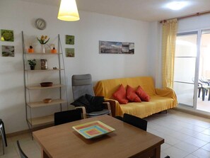 A living room with a sofa, a dining table, a chair, a bookshelf, and a window with curtains.