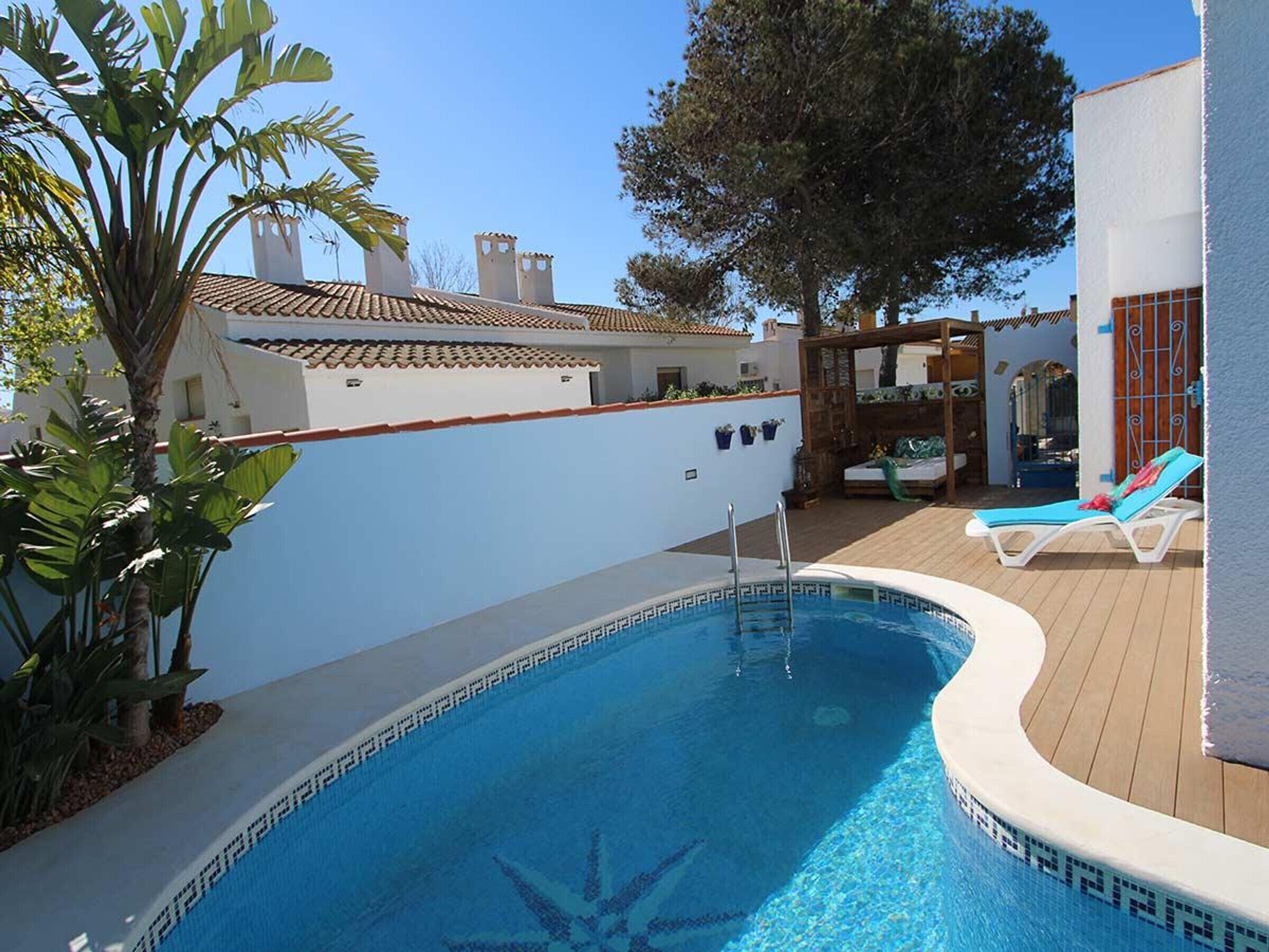 Una piscina con borde de azulejos, rodeada por un muelle de madera y un muro blanco.