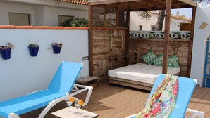 A wooden deck with two blue lounge chairs, a small table with a hat and glasses, and a bed with a wooden headboard. - Beachside Getaway: Private Pool, Free WiFi, Pet Friendly, Air Con, Near Dog Beach (Deltebre)