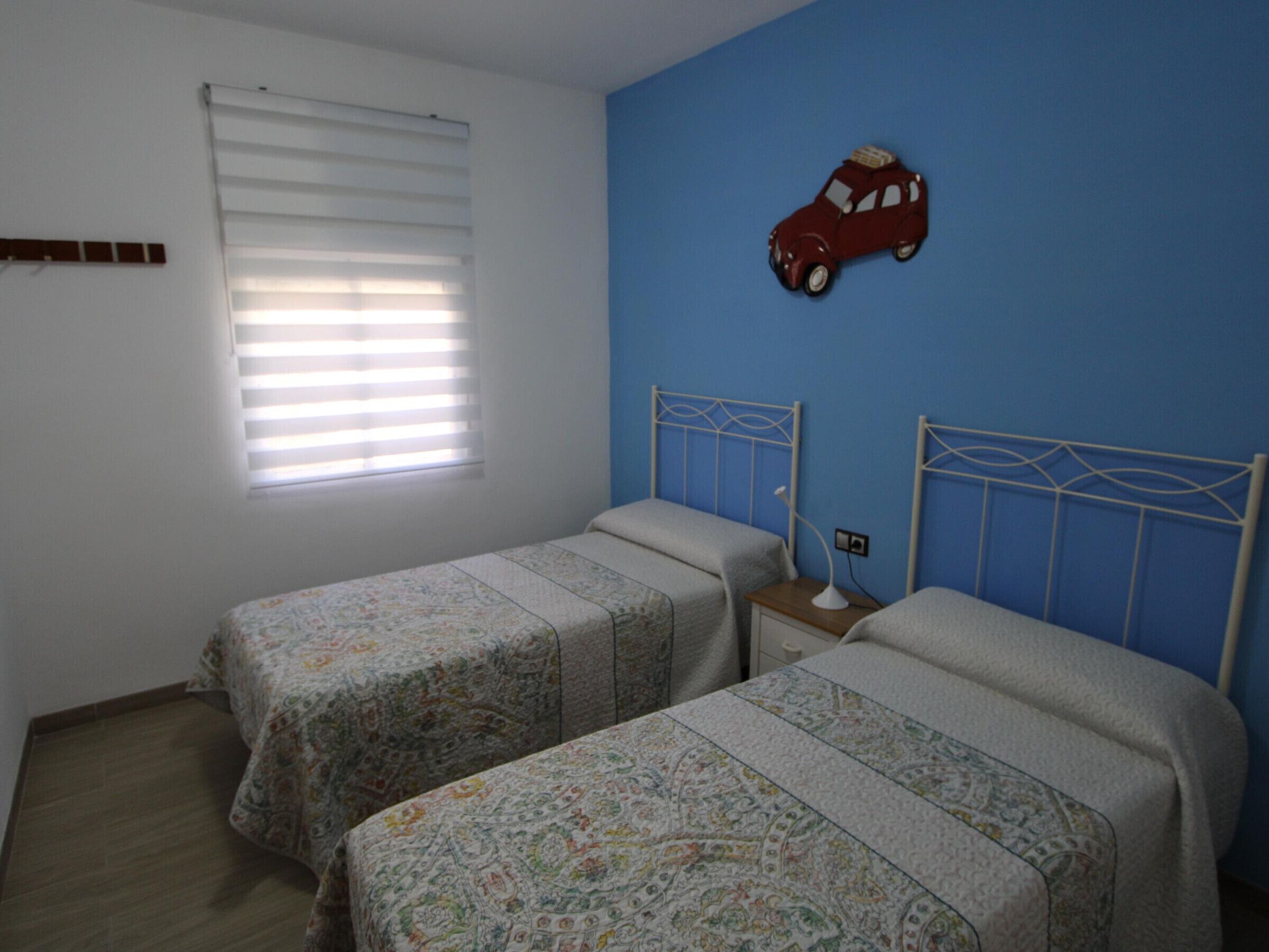 A bedroom with two single beds, a window with blinds, a wall-mounted toy car, and a bedside table with a lamp.