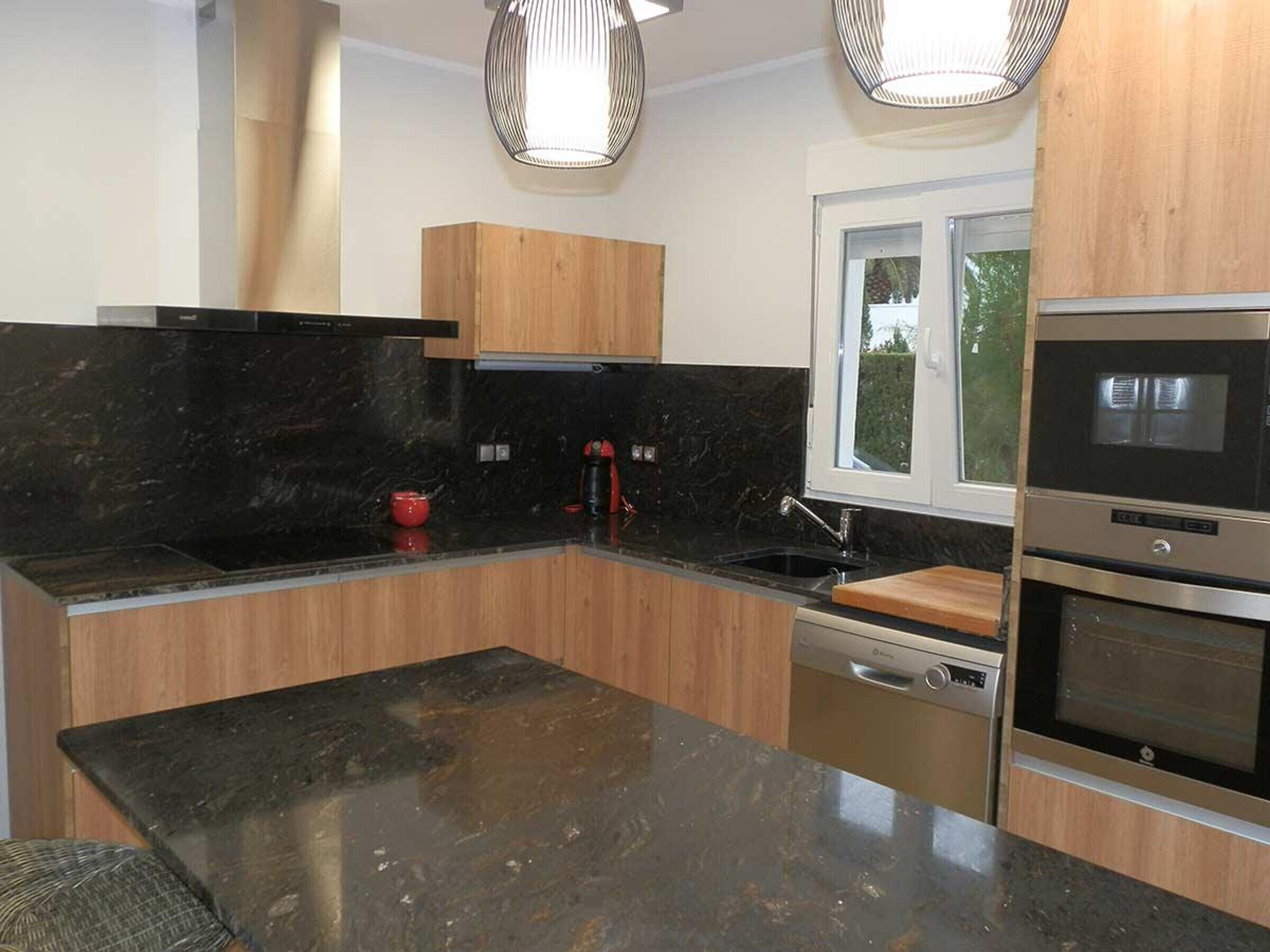 A modern kitchen with black countertops, wooden cabinets, stainless steel appliances, and pendant lighting.