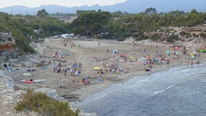 Ein Strand mit badenden und sonnenden Menschen, umgeben von felsigem Gelände und Bäumen.
