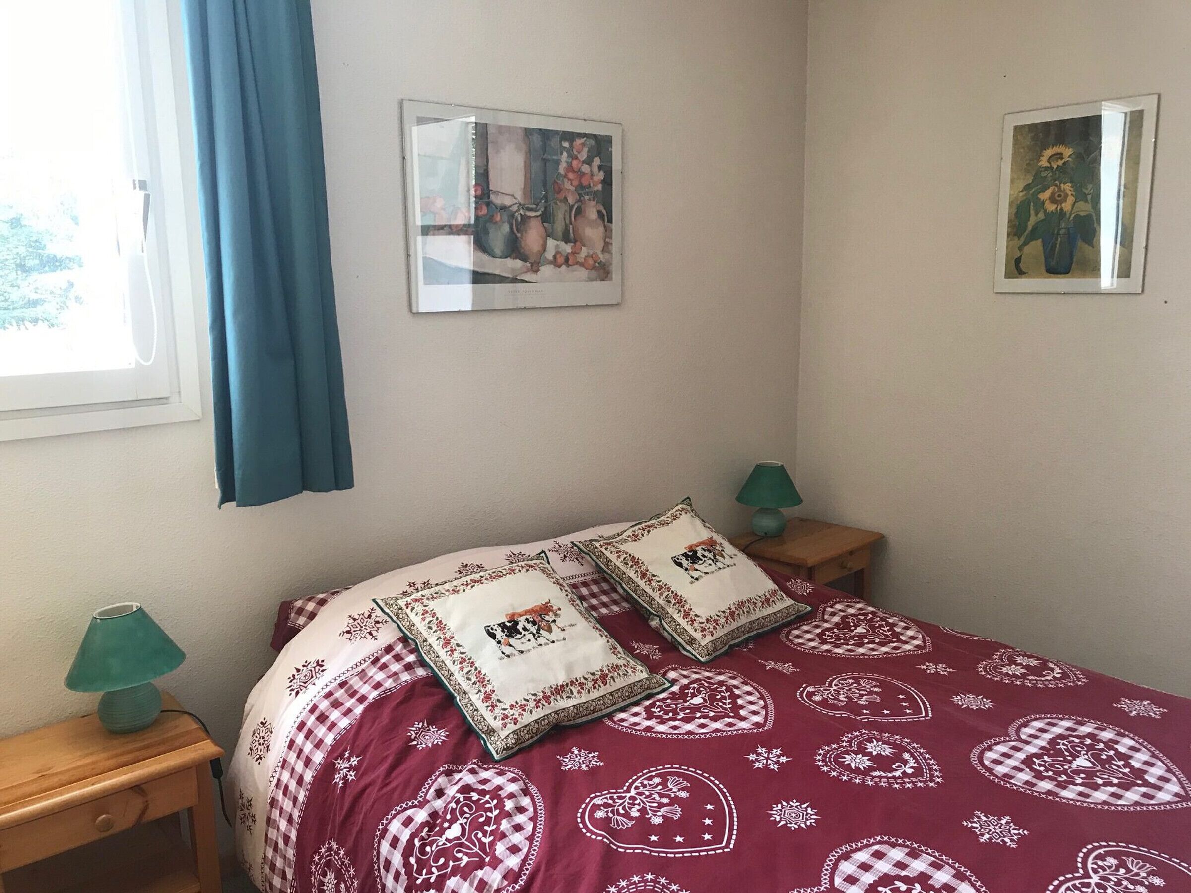 Un dormitorio con una cama, dos almohadas, una mesita de noche con una lámpara y dos cuadros enmarcados en la pared.