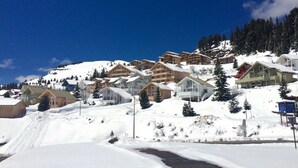 Un poble de muntanya cobert de neu amb edificis d'estil xalet, un cel blau clar i una pista d'esquí al fons.