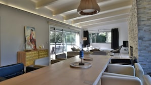 A modern living room with a long wooden table, comfortable seating, a fireplace, and large windows offering a view of the outdoors. - Maison Contemporaine Avec Piscine à Mi-chemin Entre la Plage de Sa Tuna et Begur (Begur)