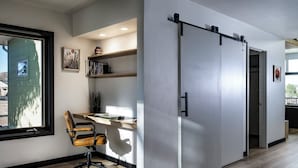 A modern office space with a sliding door, a desk with a chair, shelves with books and decorative items, and a window with a view of the outside.
