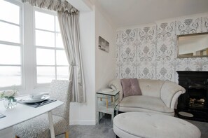 Une pièce avec une table blanche, un fauteuil à motifs, un canapé, une cheminée et une grande fenêtre avec des rideaux.