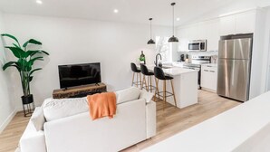 Een moderne woonkamer met een witte bank, een televisie en een plant. Het heeft een keukenhoek met een koelkast, magnetron en barstoelen.