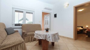 A room with a large window, a sofa, a table with a floral tablecloth, and a door leading to another room.