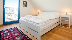 A bedroom with a white bed, a small bedside table with a lamp, a window with a blue frame, and a patterned rug. - Gorgeous home in Rowy with house a panoramic view (Rowy)