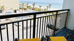 Un balcon avec vue sur la plage, un espace de stationnement et un bâtiment en arrière-plan.