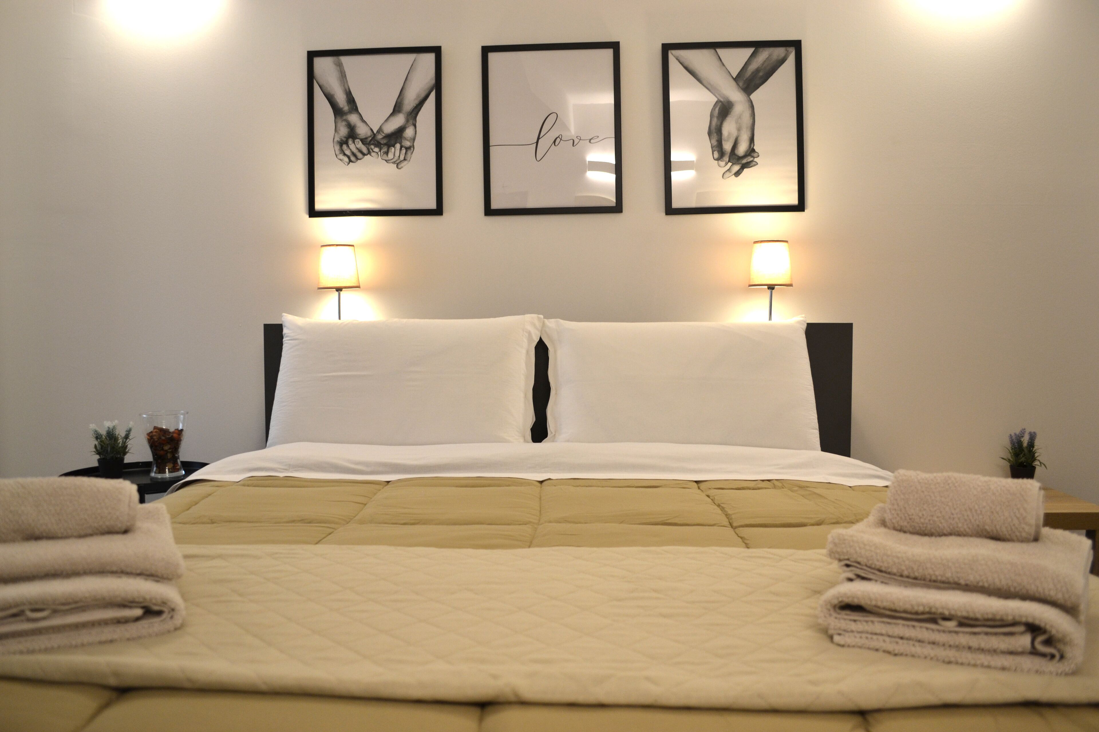 Un lit bien fait, recouvert d’une courtepointe beige, avec deux oreillers blancs et une paire de serviettes pliées de chaque côté. Trois œuvres d’art encadrées sont accrochées au-dessus du lit, et deux lampes de chevet sont placées de chaque côté.
