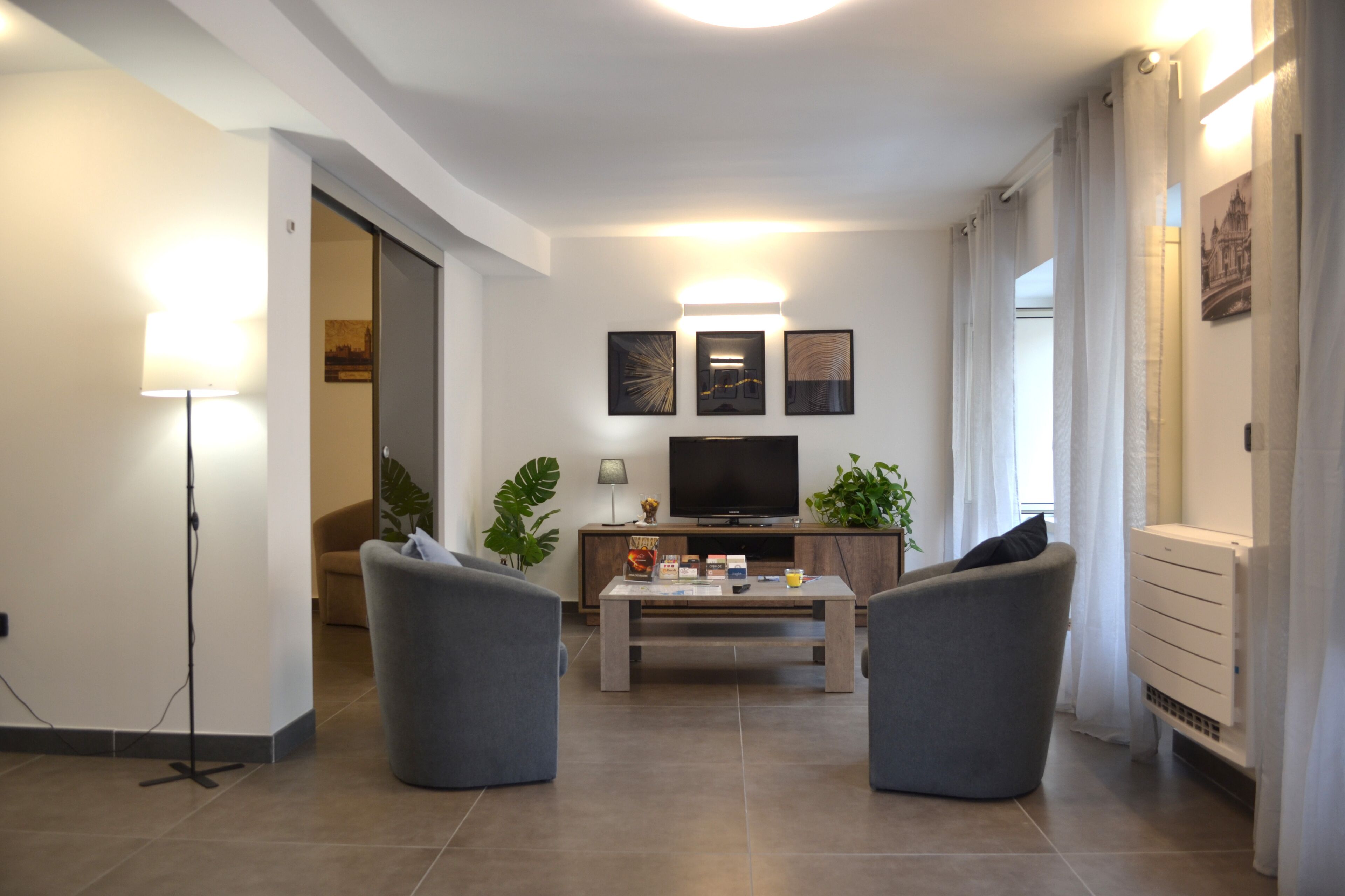 Ein modernes Wohnzimmer mit einem Flachbildfernseher, zwei grauen Sesseln, einem Couchtisch und einer Stehlampe. Darüber an der Wand befinden sich drei gerahmte Kunstwerke.