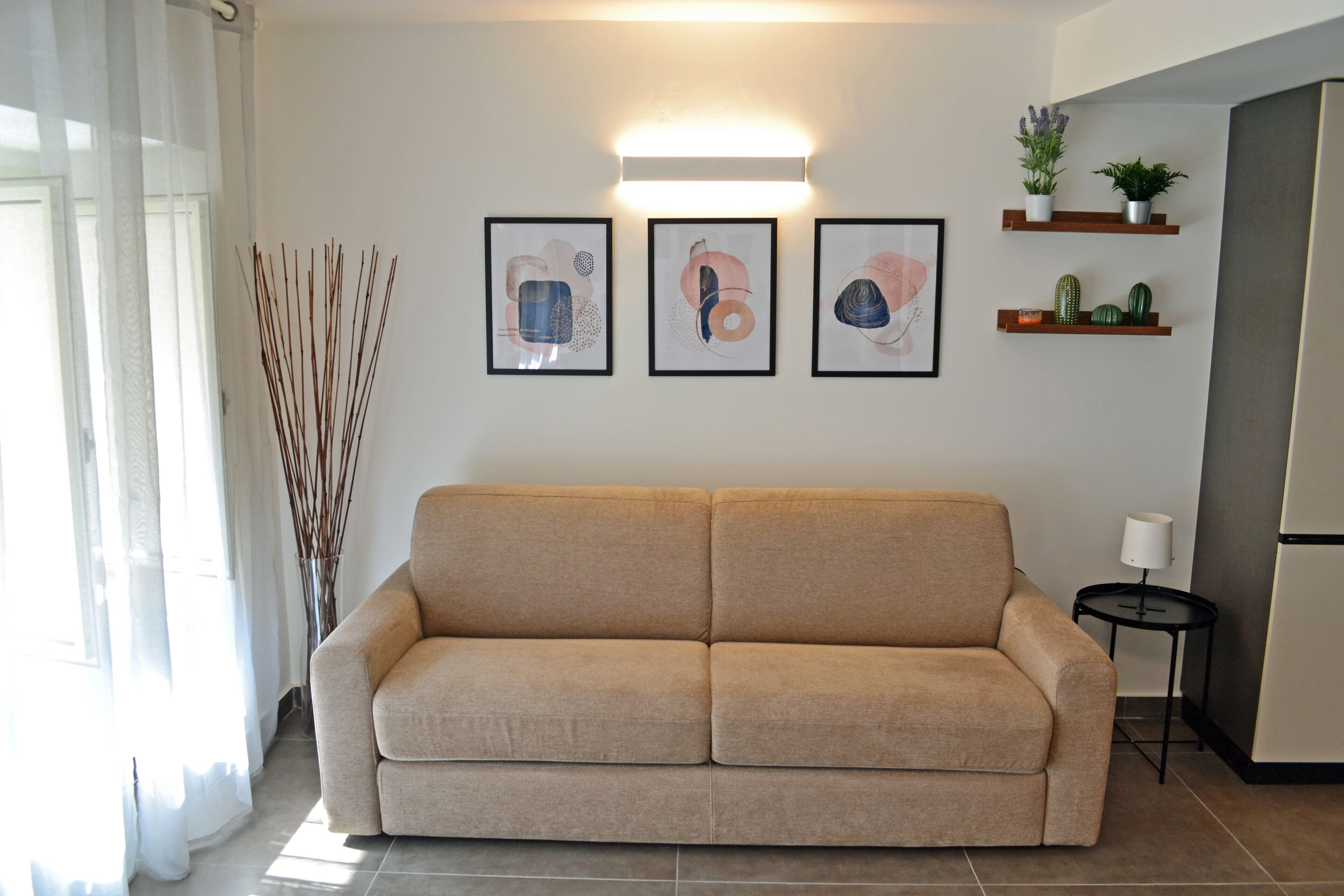 A living room with a beige sofa, three framed abstract artworks, a small table with a lamp, and a refrigerator.