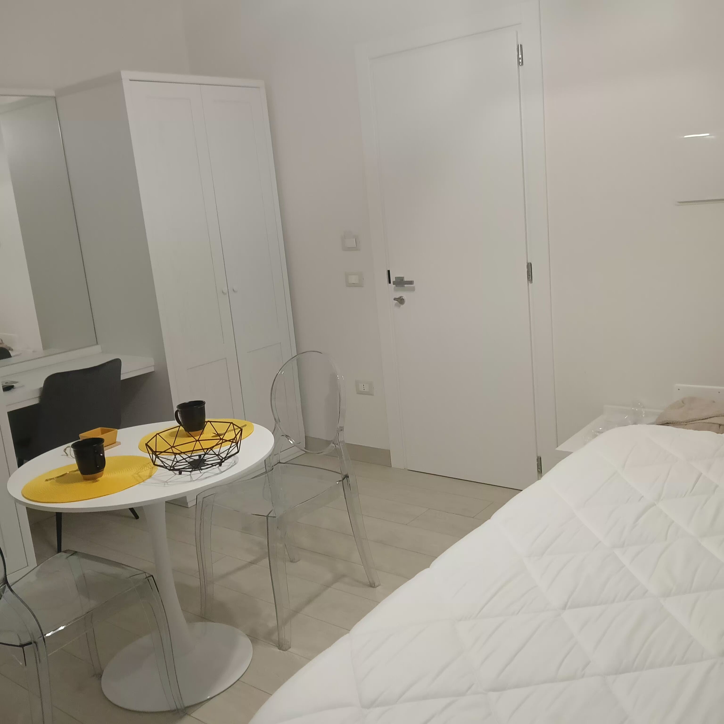 A hotel room with a bed, a small round table with a yellow placemat, a black mug, and a geometric-patterned tray. There is a white wardrobe, a door, and a mirror on the wall.
