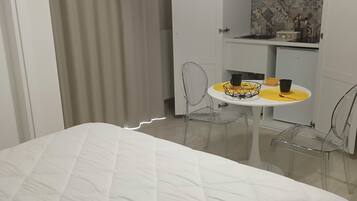 Habitación de hotel con cama, una mesita pequeña con camino de mesa amarillo, dos sillas, kitchenette y baño visible tras una cortina.