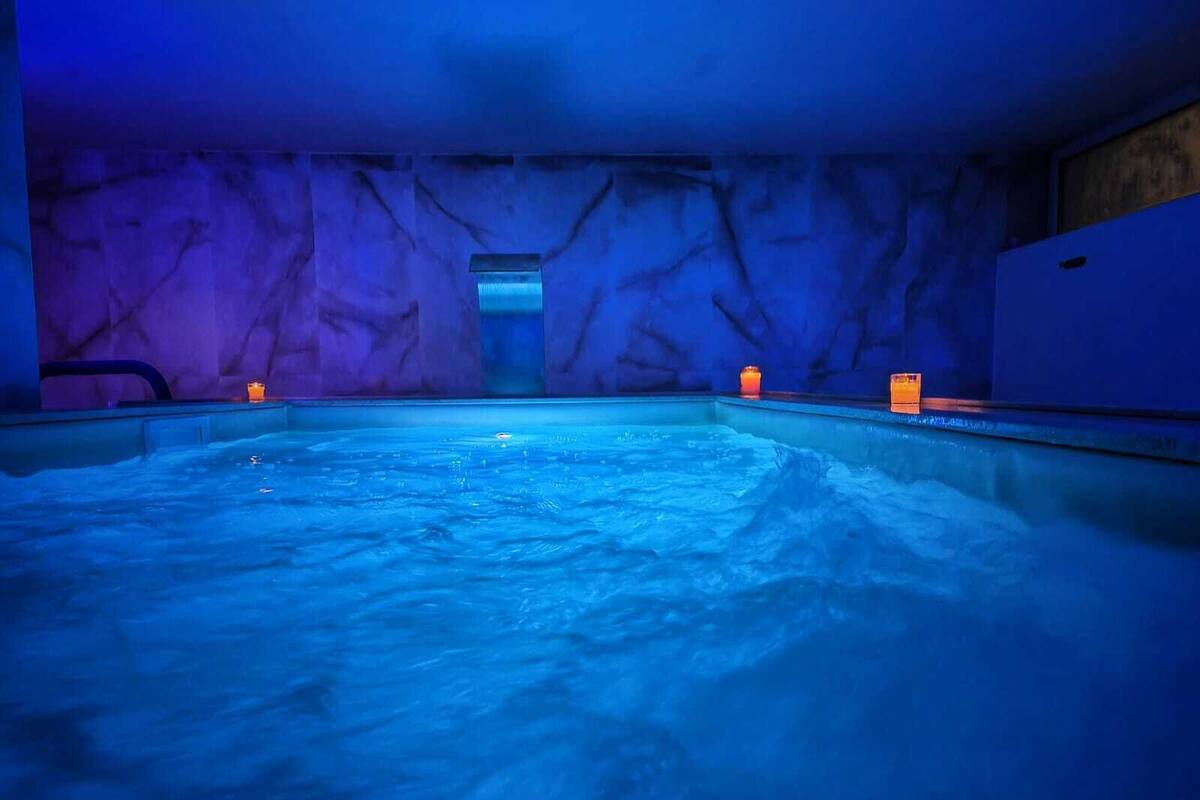 A large indoor swimming pool with a blue-lit ambiance, featuring a stone wall backdrop and two orange candles on the poolside.