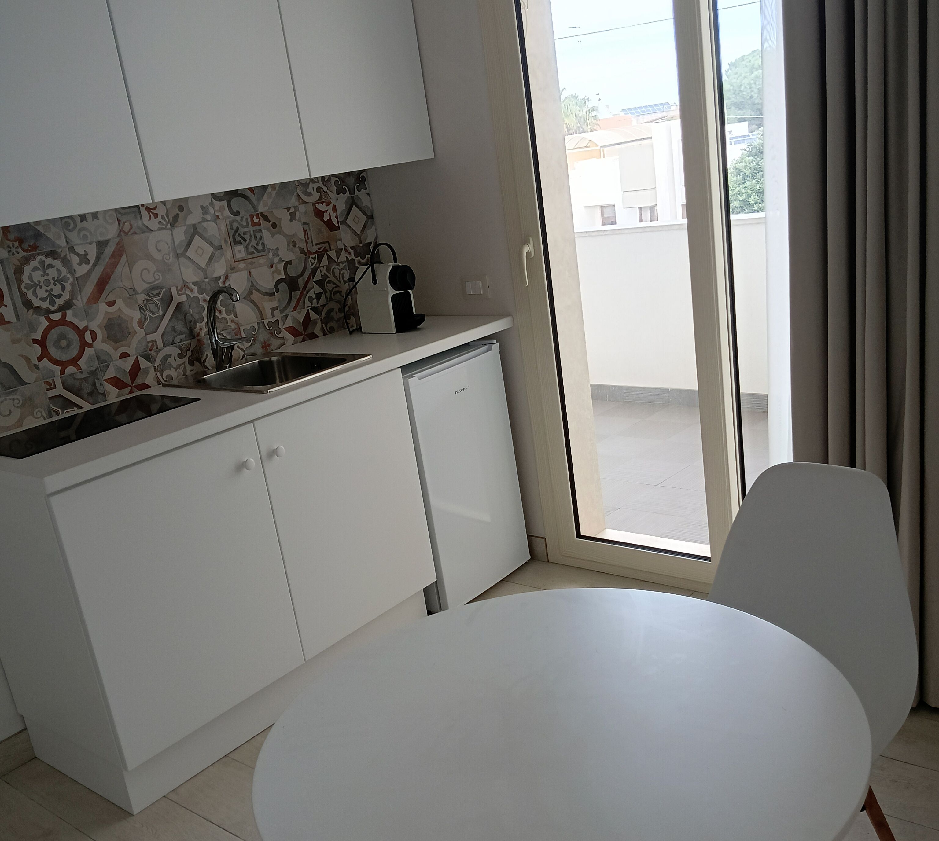 A modern kitchen with white cabinets, a patterned backsplash, and a built-in dishwasher. There is a dining table with chairs and a view of the outside through a sliding glass door.