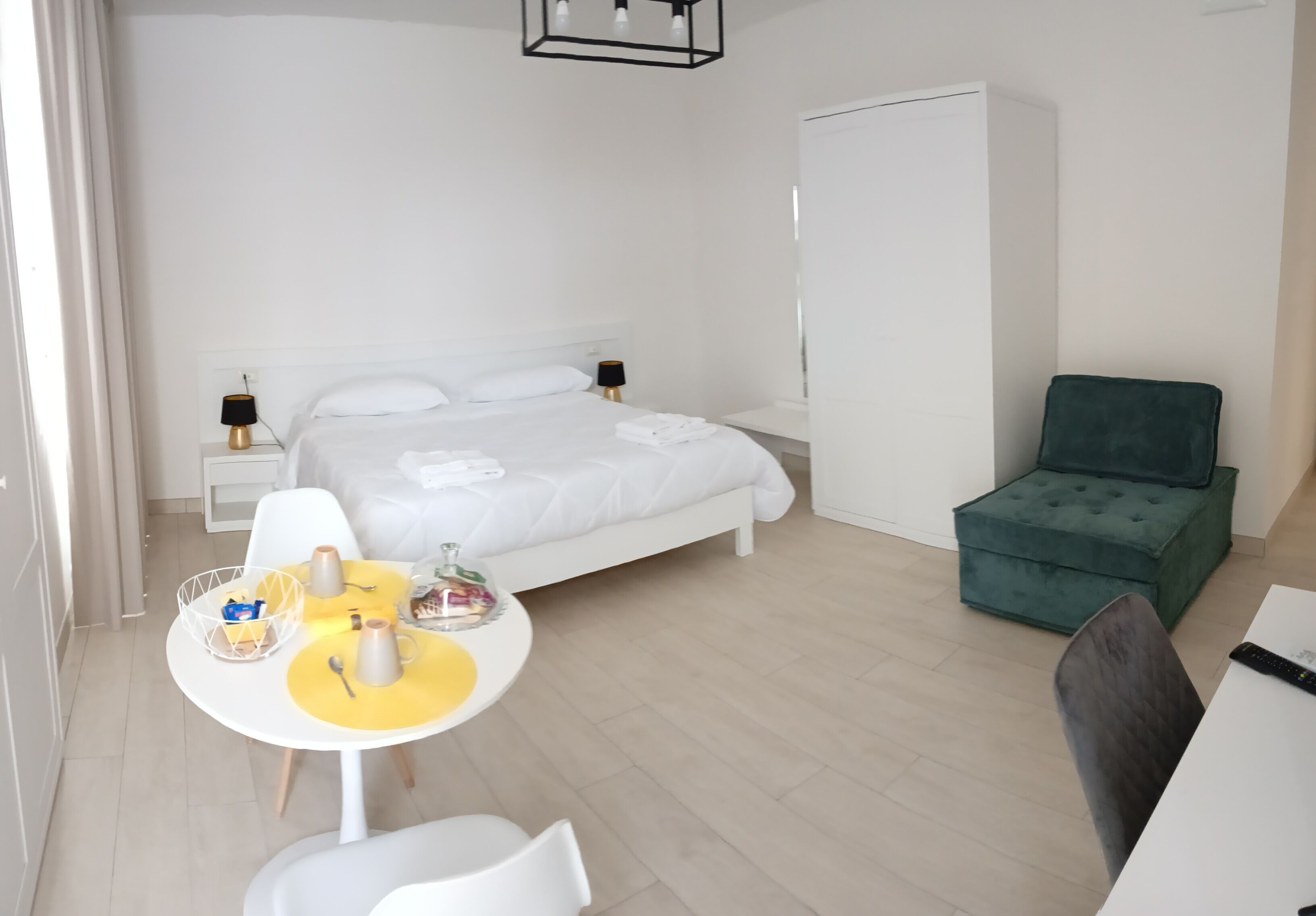 Ein modernes Hotelzimmer mit einem Bett, Nachttischen, einem grünen Sessel, einem Esstisch mit gelben Platzsets und einem weißen Kleiderschrank.