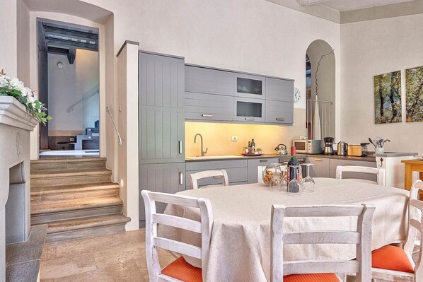 A modern kitchen with grey cabinets, a central island, and a dining area with white chairs and orange cushions. - Villa Church with private pool - My Rental Homes (Cortona)