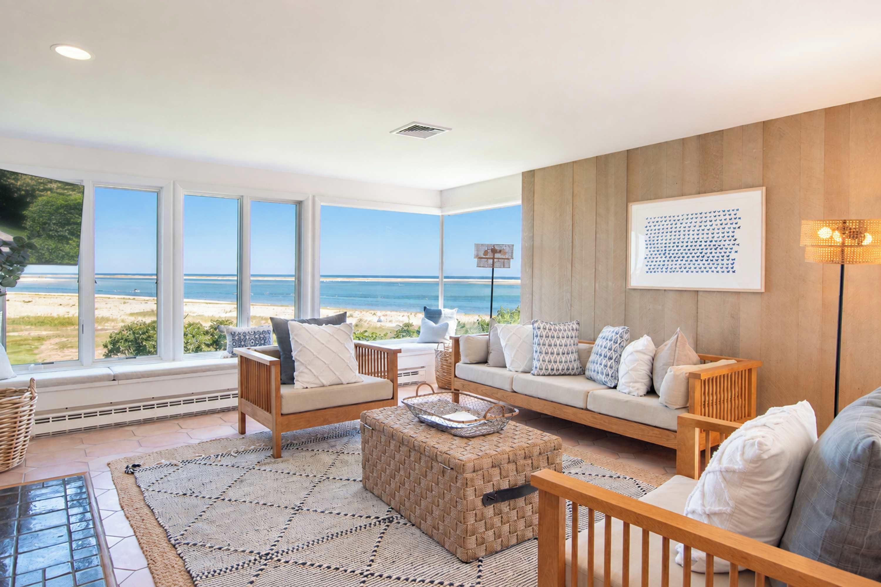 A spacious living room with a large window overlooking a beach, a wooden wall, and a framed artwork.