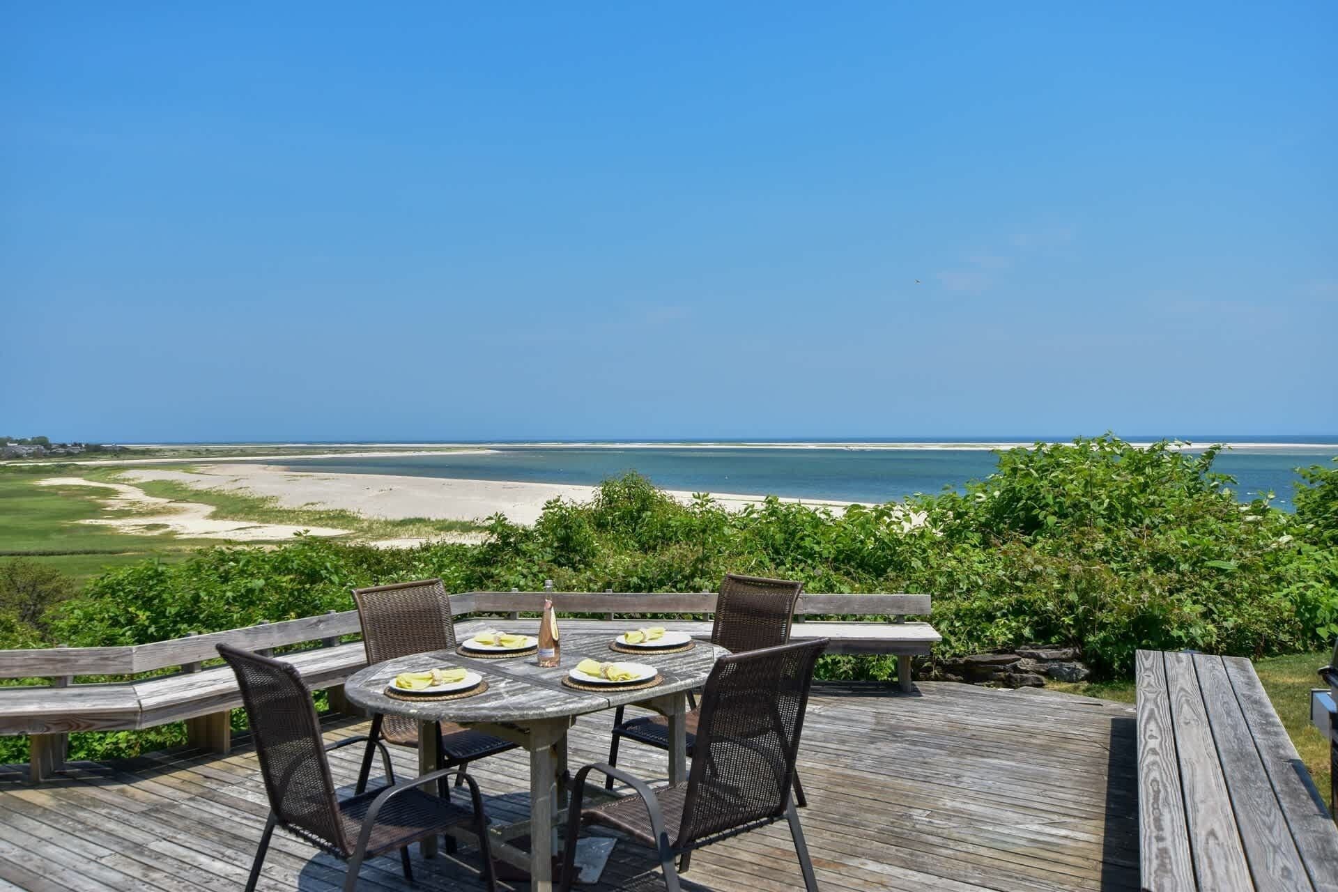 Een houten terras met een tafel en stoelen, uitkijkend op een strand.