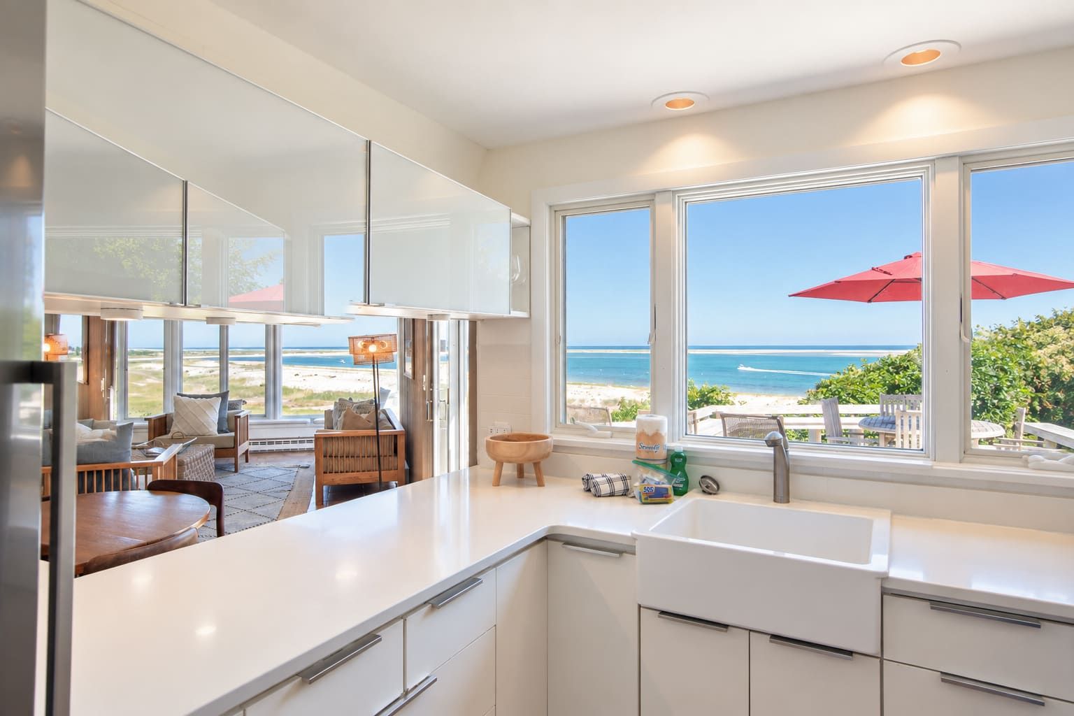 Une cuisine moderne dotée de grandes fenêtres donnant sur une plage, un coin repas avec vue et un bar agrémenté d’un parasol rouge.