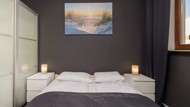 Un dormitorio con cama, dos mesitas de noche con lámparas, un cuadro de un atardecer sobre el agua y una ventana con cortinas.